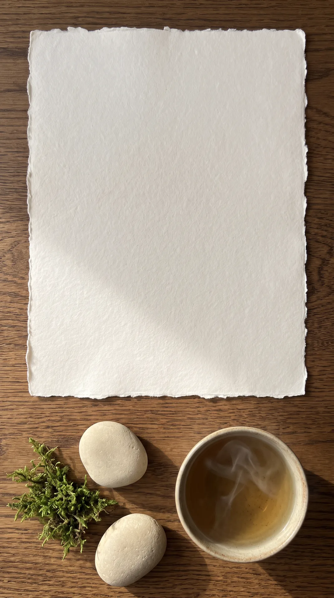 Eikenhouten tafel met een groot vel handgeschept papier, dampende keramische theekom, twee gladde rivierstenen en groen mos